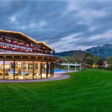 A modern hotel building with many windows, surrounded by a green meadow and mountains in the background. The sky is overcast and it is slightly twilight.