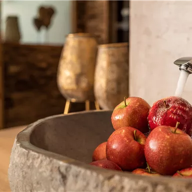 A stone bowl with fresh apples is on a wooden table. In the background, water flows from a modern faucet.