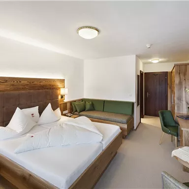 A cozy hotel room with a large bed, a corner for relaxing, and wooden details. The room is bright and decorated in a modern style.