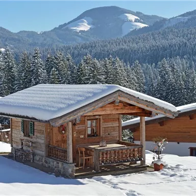 A cozy wooden cabin in the snow, surrounded by snow-covered mountains and forests. The clear sky and winter landscape create a peaceful atmosphere.