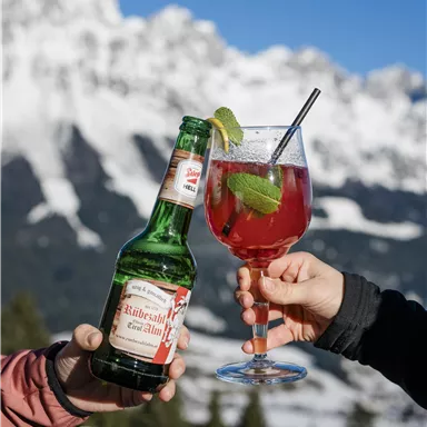 Ein Bier und ein Cocktail werden in den Bergen angestoßen. Im Hintergrund sind schneebedeckte Berge und blauer Himmel zu sehen.