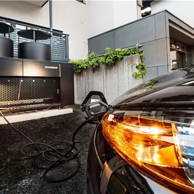 A modern electric car charging station next to a stylish vehicle. In the background, some buildings and plants are visible.