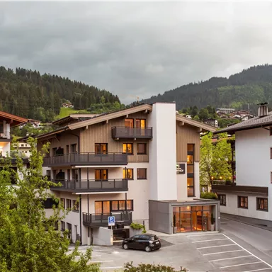 A modern building with several floors and balconies, surrounded by trees and mountains. The sky is overcast and the surroundings seem tranquil.