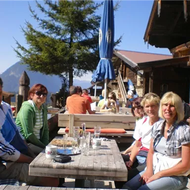 A group of five people is sitting at a wooden table in a mountain hut. In the background, there are more guests and trees visible, surrounding the beautiful mountain landscape.