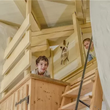 Zwei Kinder schauen von einer Holztreppe in einem hellen, freundlichen Raum. Die Atmosphäre ist einladend und verspielt.