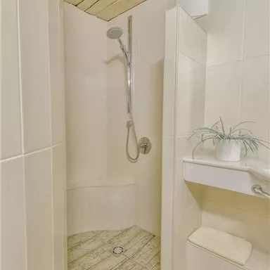 Eine moderne Dusche mit hellem Fliesenboden und Holzdecke. Ein kleiner Ablagetisch mit einer Pflanze und Handtuch ist ebenfalls sichtbar.