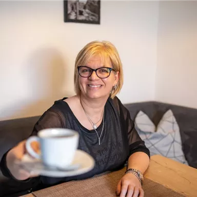 Eine lächelnde Frau sitzt an einem Tisch und hält eine Kaffeetasse hoch. Im Hintergrund sind eine gemütliche Sitzecke und dekorative Kissen zu sehen.