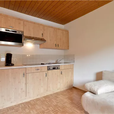 A modern kitchen unit with wooden cabinets and a stove. Next to it is a cozy sofa in a bright room.