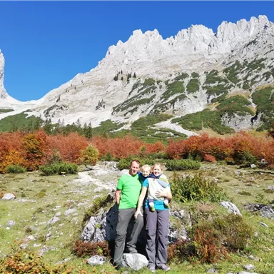 A family poses in front of an impressive mountain backdrop with colorful autumn foliage. The sky is clear and blue, highlighting the natural beauty of the landscape.