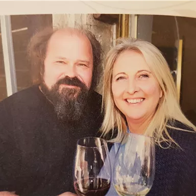 Ein Paar posiert lächelnd vor einer Wand. Beide halten ein Weinglas in der Hand.