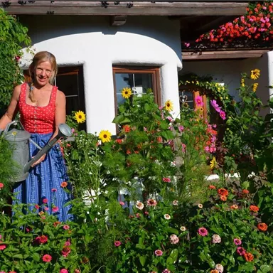 Eine Frau gießt Blumen in einem bunten Garten vor einem gemütlichen Haus. Der Garten ist voller bunten Blumen und strahlendem Sonnenlicht.