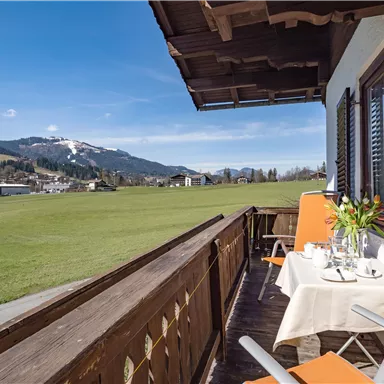 Ein Balkon mit Blick auf eine grüne Wiese und die Berge im Hintergrund. Auf dem Tisch stehen Blumen und die Stühle sind orange.