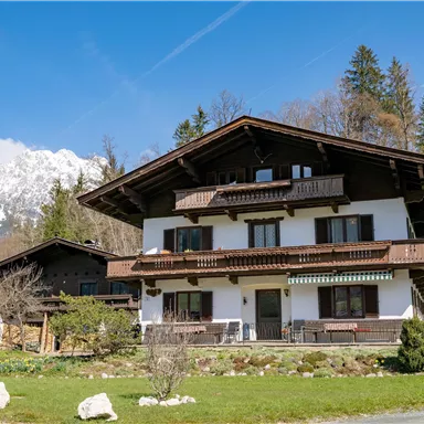 Ein charmantes Haus im alpinen Stil, umgeben von grünen Wiesen und Bergen im Hintergrund. Der Himmel ist blau und klar.