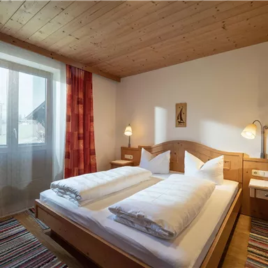 Ein gemütliches Zimmer mit einem Doppelbett und zwei Kopfkissen. Das Zimmer hat große Fenster mit einen Blick nach draußen und eine Holzdecke.