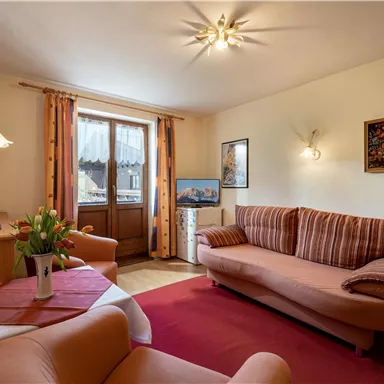 Ein gemütliches Wohnzimmer mit einem roten Teppich und bequemen Sofas. Es gibt Fenster mit Vorhängen und einen Fernseher auf einem kleinen Tisch.