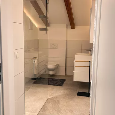 Ein modernes Badezimmer mit hellen Fliesen und Holzbeamten. Im Hintergrund ist eine Dusche und ein Waschtisch zu sehen.