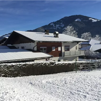 Ein verschneites Haus vor einer malerischen Berglandschaft. Der Garten ist mit Schnee bedeckt und der Himmel ist klar und blau.