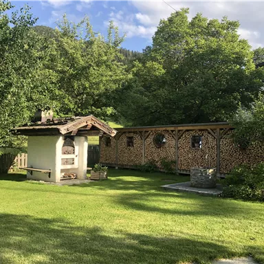 Ein schöner Garten mit einer überdachten Sitzecke und einer Holzstoßwand. Im Hintergrund sind Bäume und eine ländliche Umgebung zu sehen.