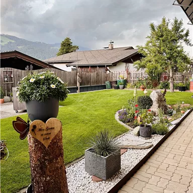 Ein gepflegter Garten mit grünem Rasen, bunten Blumen und verschiedenen Pflanzen. Im Hintergrund sind Holzhäuser und ein bewölkter Himmel zu sehen.