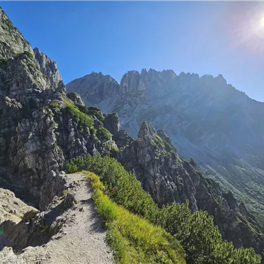 A picturesque hiking trail winds through rocky mountains under a clear blue sky. The sun shines brightly over the landscape.