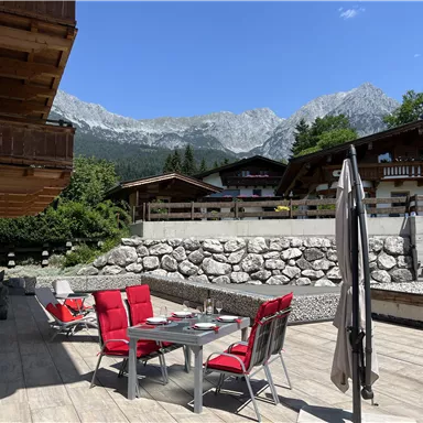 A picturesque outdoor area with a dining table and red chairs. In the background, majestic mountains and green trees can be seen.