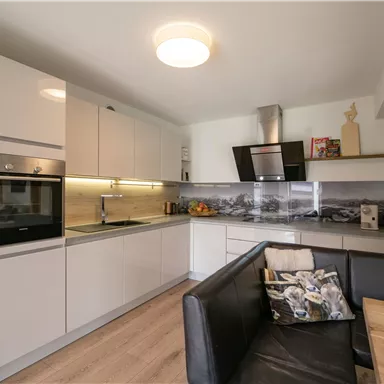 Moderne Küche mit weißen Schränken und einer eleganten Arbeitsplatte. Ein gemütlicher Wohnbereich mit einem schwarzen Sofa und einem Esstisch ist ebenfalls zu sehen.