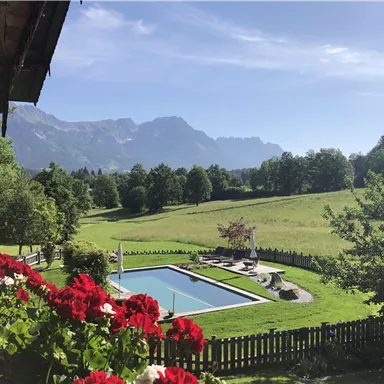 Ein schöner Blick auf einen Garten mit einem Pool, umgeben von grünen Wiesen und Bäumen. Im Hintergrund sind majestätische Berge und ein klarer blauer Himmel zu sehen.