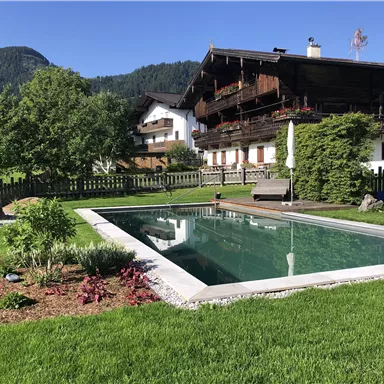 Ein gepflegter Garten mit einem Pool und einer Holzterrasse. Im Hintergrund sind traditionelle Häuser und grüne Berge zu sehen.