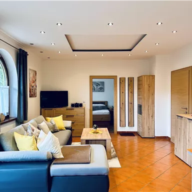 Ein modernes Wohnzimmer mit einem bequemen Sofa, Holzmöbeln und einem Fernseher. Die Fenster bieten viel Tageslicht und eine gemütliche Atmosphäre.