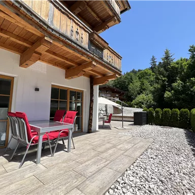 A terrace with a table and red chairs. In the background, trees and a sunny sky structure can be seen.