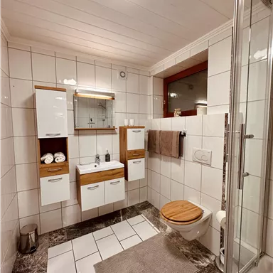 Ein modernes Badezimmer mit hellen Fliesen und Holzakzenten. Die Dusche ist durch eine Glastür abgetrennt und der Raum wirkt sauber und einladend.