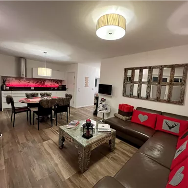 A modern living room with a cozy sofa and a dining table. The walls are light and there are decorative elements with red accents.