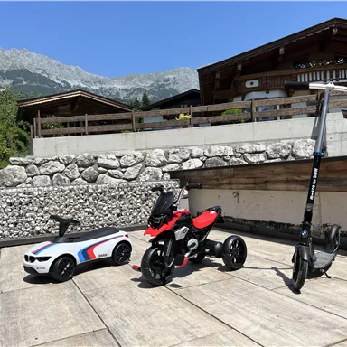 A toy car and a children's vehicle are on a terrace in nice weather. In the background, there are mountains and a modern house.