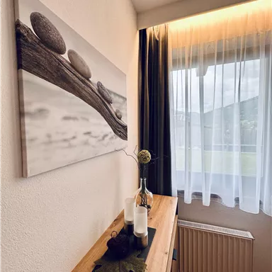 Ein gemütlicher Raum mit einem Holztisch und einer Dekoration aus Steinen und einer Vase. Das Fenster ist mit einem weißen Vorhang verziert und lässt sanftes Licht herein.
