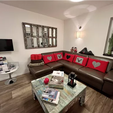 A modern living room with a brown sofa and red cushions. In the room, there is a table with magazines and a television on the wall.