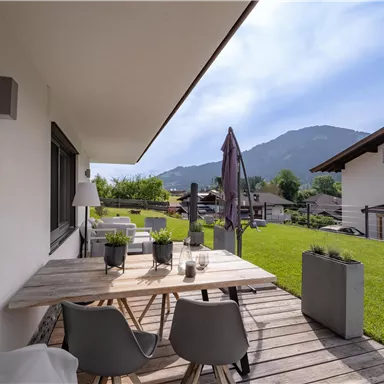 Ein gemütlicher Außenbereich mit einem Holztisch und modernen Stühlen. Im Hintergrund sind grüne Wiesen und Berge zu sehen.