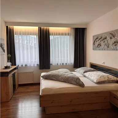 Ein gemütliches Schlafzimmer mit einem großen Bett und Holzfußboden. Große Fenster mit Vorhängen lassen viel Licht herein.