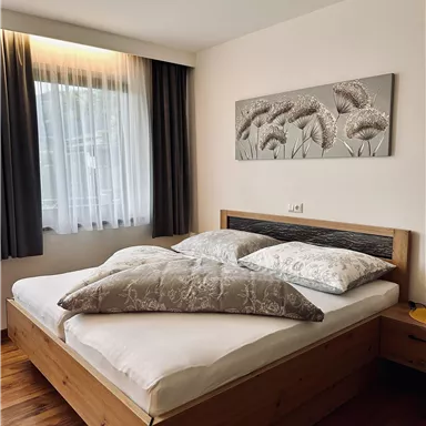 Ein gemütliches Schlafzimmer mit einem großen Doppelbett und zwei Kissen. Sanfte Farben und ein großes Fenster sorgen für ein helles Ambiente.