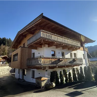 A modern house in alpine style with wooden cladding and several balconies. The surroundings are surrounded by green trees and mountains.