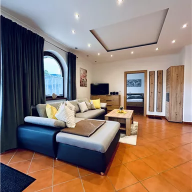 Ein modernes Wohnzimmer mit einem schwarzen Sofa, bunten Kissen und einem großen Fenster. Der Boden ist mit terrakottafarbigen Fliesen ausgelegt und es gibt einen kleinen Holztisch.