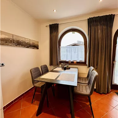 Ein modernes Esszimmer mit einem Tisch und vier Stühlen. Große Fenster mit Vorhängen sorgen für viel Licht.