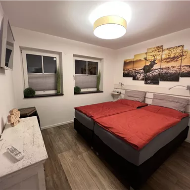 A modern bedroom with a double bed and red bed covers. The walls are adorned with decorative pictures, and there are windows that let in daylight.