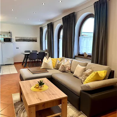 Ein gemütliches Wohnzimmer mit einem bequemen Sofa und einem Holztisch. Große Fenster sorgen für viel Licht und eine einladende Atmosphäre.