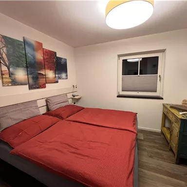 A cozy bedroom with a double bed and red bedspreads. Colorful pictures hang on the wall, and the room is bright and modernly furnished.