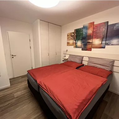 A cozy bedroom with a large double bed and red bedcovers. Colorful pictures hang on the wall, and there is a small table and a wardrobe.