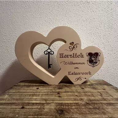 A heart-shaped wooden decoration with the inscription "Welcome to the Cat Workshop" and a key. The decoration sits on a wooden table and creates an inviting impression.