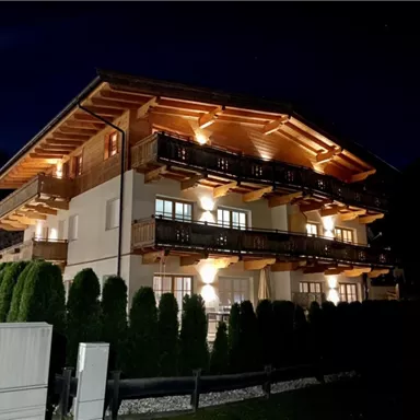 A beautiful wooden building with several balconies, illuminated at night. Surrounded by tall hedges and a tranquil ambiance.