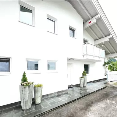 Ein modernes, weißes Haus mit großen Fenstern und einem überdachten Balkon. Auf dem Gehweg stehen zwei stilvolle Pflanzkübel.