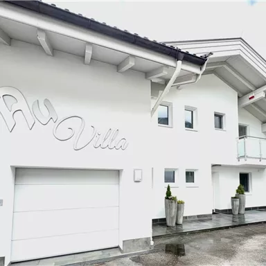 Eine moderne Villa mit einer weißen Fassade und großem Garagentor. Der Eingang ist mit Pflanzen dekoriert und das Dach hat einen klassischen Überstand.