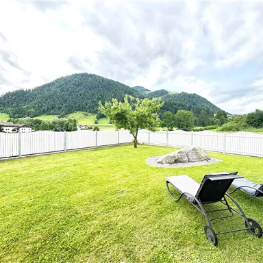 Eine grüne Wiese mit zwei Liegen und Blick auf sanfte Hügel. Im Hintergrund sind Berge und ein blauer Himmel zu sehen.